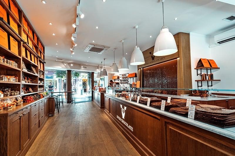 Bright Swiss chocolate shop interior with warm wood counters, orange gift boxes, and recessed downlight illumination.